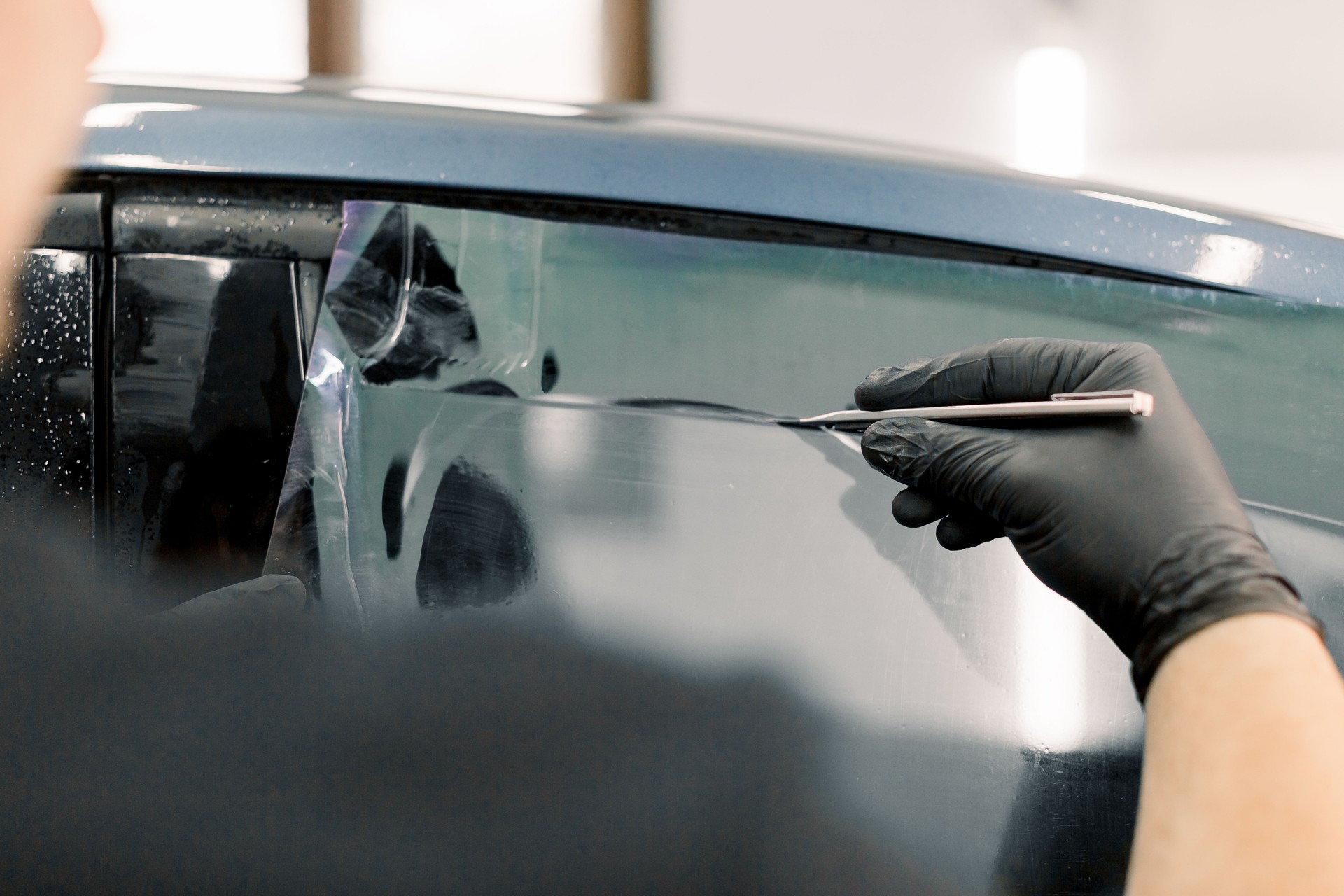 Cropped image of hands of worker in garage tinting a car window with tinted foil or film, holding special blade or knife to cut the film. Car detailing workshop, tinting windows